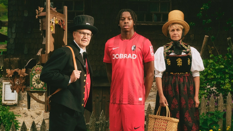 Ein SC Spieler steht vor dem Hof, daneben 2 historische Kaufleute mit Kuckucksuhren